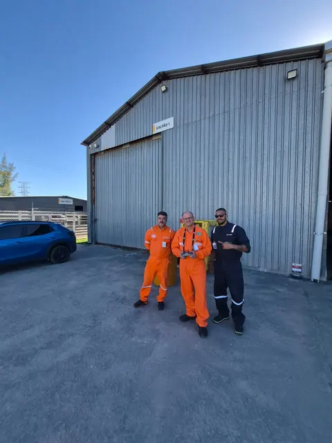 Instrutor e dois alunos de macacão laranja em frente a um hangar